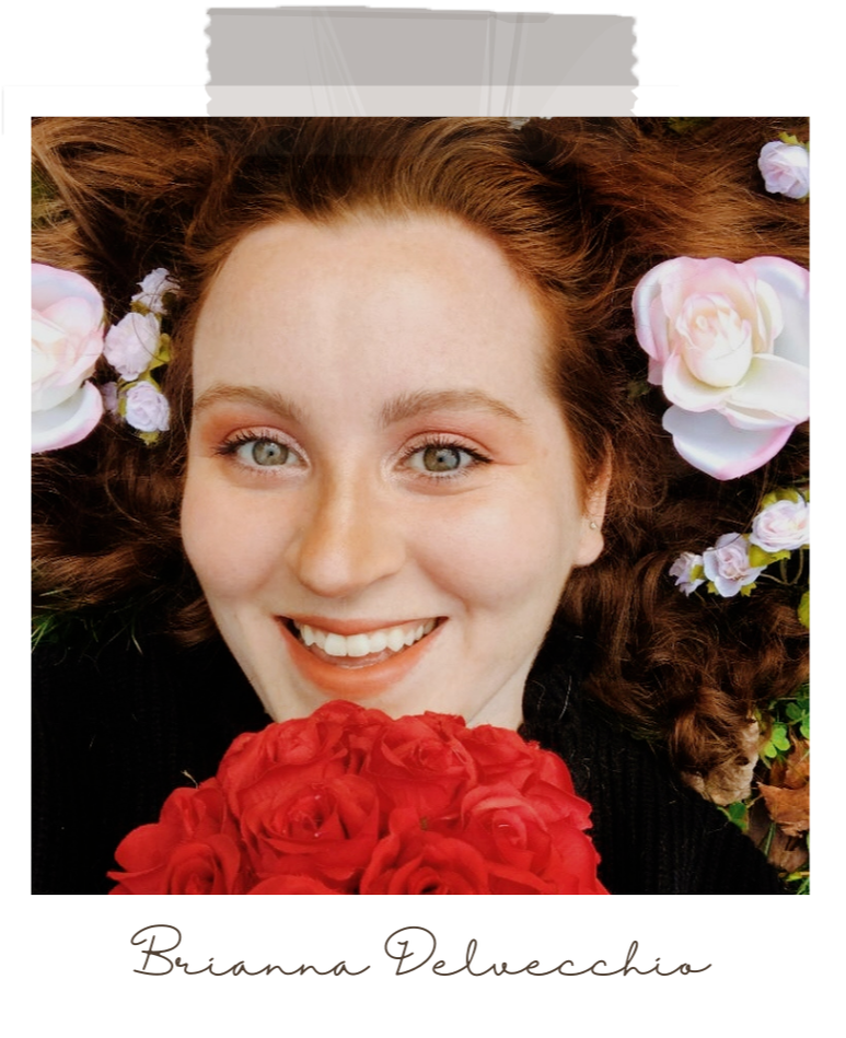 Brianna Delvecchio is laying down, with flowers scattered along her spread out hair. She is smiling at the camera, while holding a bouquet of roses.