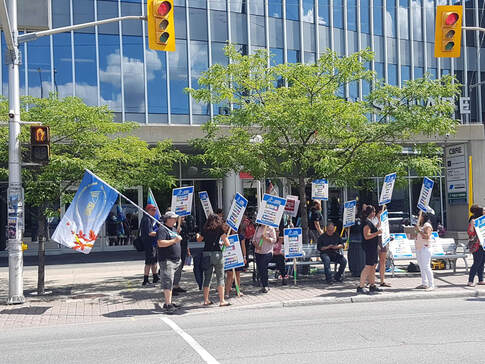 An OPSEU Rally: a large group of people (10-15) stand by a crosswalk in a city with flags and signs. 